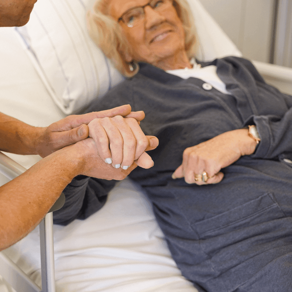 Äldre kvinna som ligger i säng och håller hand med en annan person