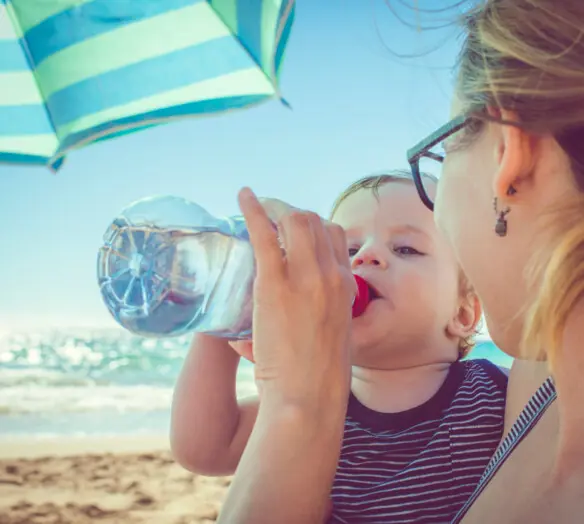 Litet barn på stranden dricker vatten ur flaska som kvinna håller i. 